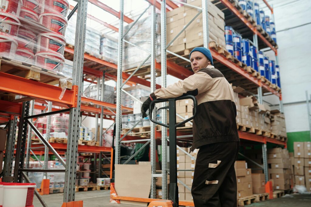 Warehouse worker operating a pallet jack among stacked boxes and metal shelves in an industrial setting.
